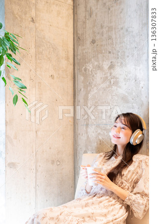 ソファーに座りヘッドホンで音楽を聴きながらコーヒーを飲んでくつろいでいる若い女性 136307833