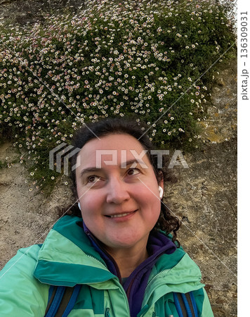 Smiling solo female pilgrim taking selfie near stone wall covered with small white wildflowers along Camino de Santiago route 136309031