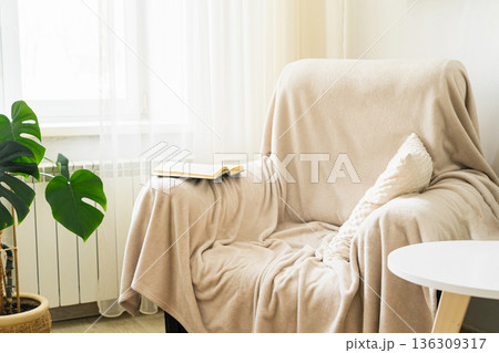 Armchair draped with a soft blanket and an open book by the window create a cozy nook for reading and relaxing 136309317