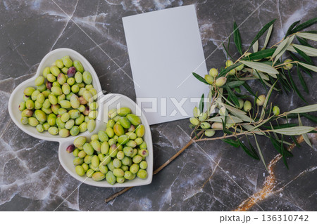 Freshly harvested green olives in heart shaped white plate with olive tree branch, blank paper for copy space, top view Mediterranean food concept Freshly harvested green olives in heart shaped white plate with olive tree branch, blank paper for copy space, top view Mediterranean food concept 136310742