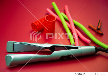 Top view of a green hair straightener with a twisted ceramic canvas, next to which there are curlers of different shapes and sizes on a pink background. The concept of styling, hairstyle, curls. High Top view of a green hair straightener with a twisted ceramic canvas, next to which there are curlers of different shapes and sizes on a pink background. The concept of styling, hairstyle, curls. High 136311305