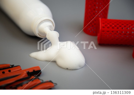 Side view of a bottle of foam for long-term hair styling, white foam poured out of it, next to it are pink curlers of different shapes and sizes on a gray background. The concept of styling, hairstyle Side view of a bottle of foam for long-term hair styling, white foam poured out of it, next to it are pink curlers of different shapes and sizes on a gray background. The concept of styling, hairstyle 136311309