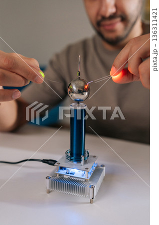 Vertical shot of scientist holding two leds near mini tesla coil, observing electrostatic discharge and electrical sparks illuminating light-emitting diodes during physics experiment, closeup. 136311421