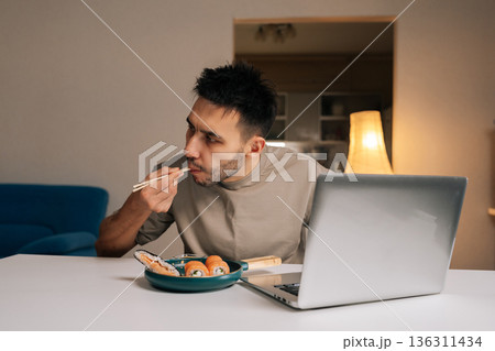 Bearded young man eating Asian delivery food with chopsticks while using laptop at home, combining dinner and work. Concept of modern multitasking lifestyle, domestic comfort and remote activity. Bearded young man eating Asian delivery food with chopsticks while using laptop at home, combining dinner and work. Concept of modern multitasking lifestyle, domestic comfort and remote activity. 136311434