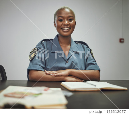 Happy, smile and portrait of a female police officer sitting by a desk in the office writing reports. Confidence, happiness and African woman security guard with a positive mindset in the workplace. Happy, smile and portrait of a female police officer sitting by a desk in the office writing reports. Confidence, happiness and African woman security guard with a positive mindset in the workplace. 136312077