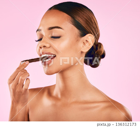 Woman is eating chocolate, health and beauty with candy, skincare and wellness on pink background. Female model with sweets snack, cacao dessert and glow with makeup and cosmetic shine in studio Woman is eating chocolate, health and beauty with candy, skincare and wellness on pink background. Female model with sweets snack, cacao dessert and glow with makeup and cosmetic shine in studio 136312173