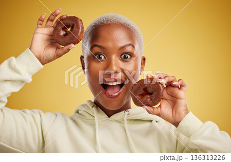 Portrait, chocolate and donut with a black woman in studio on a gold background for candy or unhealthy eating. Smile, food and baking with a happy young female person holding sweet pastry for dessert 136313226