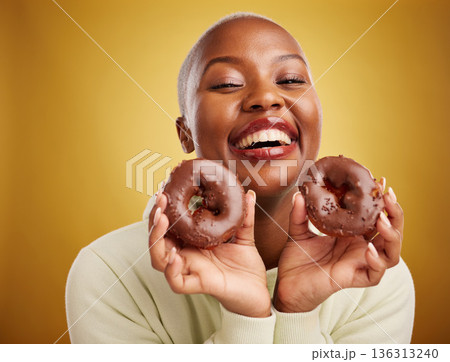 Portrait, food and donut with a black woman in studio on a gold background for candy or unhealthy eating. Smile, chocolate and baking with a happy young female person holding sweet pastry for dessert 136313240