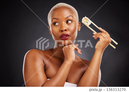 Face, thinking and woman with sushi and chopsticks in studio for healthy eating, beauty or food. Black female model with makeup idea on dark background for wellness glow, diet or seafood advertising 136313270