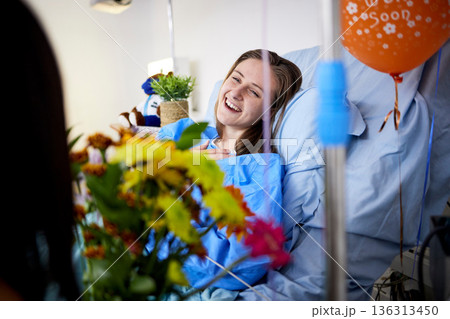 Hospital, sick patient and visitor with flowers at bed with a woman in recovery with support. Healthcare, medical insurance and person with bouquet, family and care with get well soon balloons 136313450