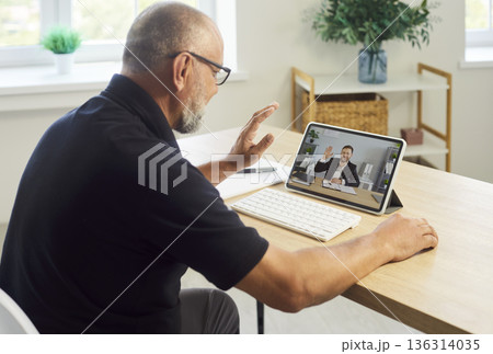 Senior man waving during online videocall on tablet at bright home office 136314035