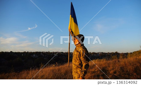 Male military in uniform lifting blue-yellow banner in honor of the victory against russian aggression at sunset. Young soldier of ukrainian army raised a waving flag of Ukraine at countryside Male military in uniform lifting blue-yellow banner in honor of the victory against russian aggression at sunset. Young soldier of ukrainian army raised a waving flag of Ukraine at countryside 136314092