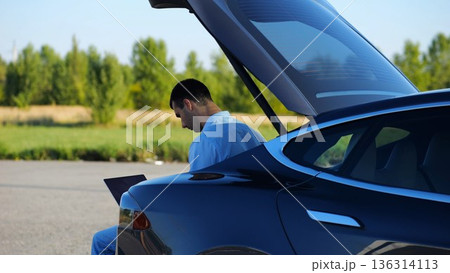 Young handsome entrepreneur works on notebook sitting in electric car trunk. Successful businessman in formal wear using computer at auto. Confident manager typing text on laptop outdoor. Slow mo 136314113