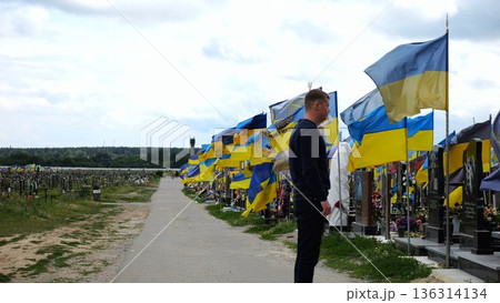Mourning man standing among countless graves of ukrainian soldiers with national banners pay tribute to fallen family member in Kharkiv. Scene reflect personal sorrow loss caused by russian aggression 136314134