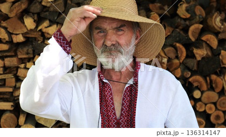 Portrait of senior ukrainian man in straw hat against the background of woodshed. Elderly serious male wearing an embroidered shirt looking into camera outdoor. Cultural heritage and ethnic roots 136314147