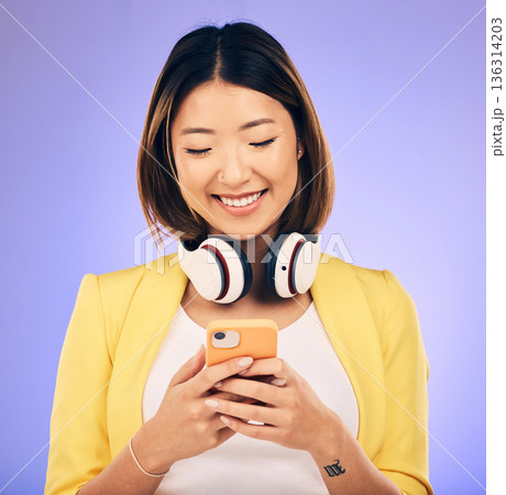Social media, phone and happy Asian woman in studio for text message, internet meme and chat. Headphones, smile and female person on smartphone listen to audio, song and track on purple background 136314203