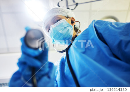 Covid, surgery and a doctor with a light in the theater for a medical procedure or inspection. Hospital, healthcare and portrait of a surgeon or clinic worker with gear for an emergency consultation 136314380