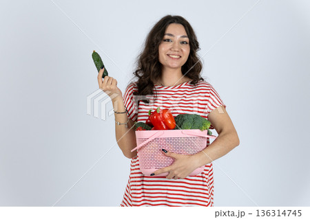 Young woman holding basket of vegetables and eating cucumber 136314745