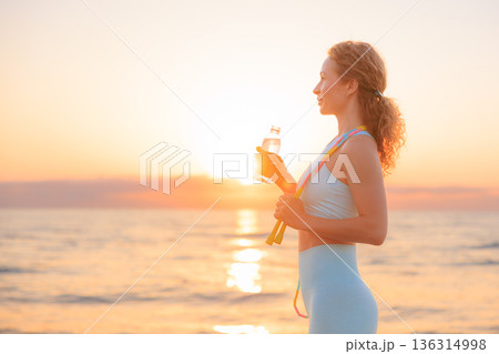 Fit woman drinking water at the beach after running outdoors in the morning. Sporty female relaxing after training. 136314998