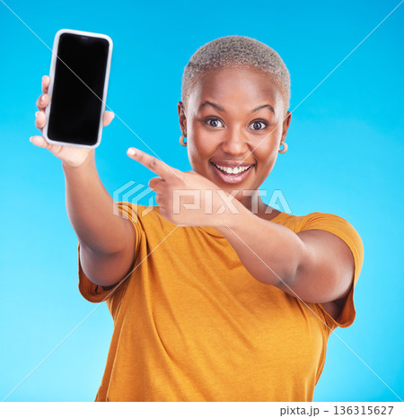 Portrait, phone screen and happy black woman point at studio advertising space, online app promo or web news. Smile, smartphone UI mockup and African person gesture at mobile deal on blue background 136315627