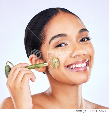 Facial roller, natural beauty and woman massage skin for wellness and glow. Portrait of a young aesthetic female model happy with dermatology, self care and face tools results on a studio background Facial roller, natural beauty and woman massage skin for wellness and glow. Portrait of a young aesthetic female model happy with dermatology, self care and face tools results on a studio background 136315874