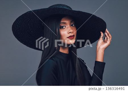 Face, fashion and serious woman with hat in studio isolated on a gray background. Portrait, style and confident female model from India with makeup cosmetics, classy clothes and elegant aesthetic. 136316491