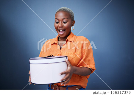 Black woman, present package and surprise in studio with excited smile, birthday and blue background. African girl, happy and wow with box, gift and fashion for celebration, winner and birthday party 136317379