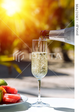 Pouring a champagne into flute glass, amazing fizz and bubbles in sparkling wine, table served with fruit plate at outdoor restaurant  on sunset with sun beams 136317496
