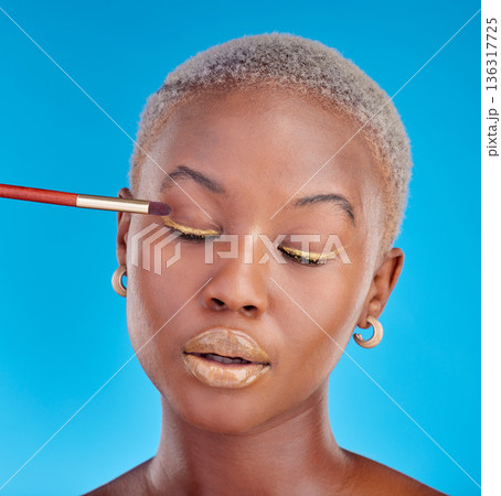 Face, makeup and eyeshadow with a black woman on a blue background in studio to apply a beauty product. Cosmetics, gold color on a brush and a young female model eyes closed for style or glamour Face, makeup and eyeshadow with a black woman on a blue background in studio to apply a beauty product. Cosmetics, gold color on a brush and a young female model eyes closed for style or glamour 136317725