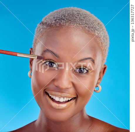 Portrait, makeup and eyeshadow with a black woman on a blue background in studio to apply a beauty product. Face, gold color on a brush and a happy young female model posing for style or glamour Portrait, makeup and eyeshadow with a black woman on a blue background in studio to apply a beauty product. Face, gold color on a brush and a happy young female model posing for style or glamour 136317727