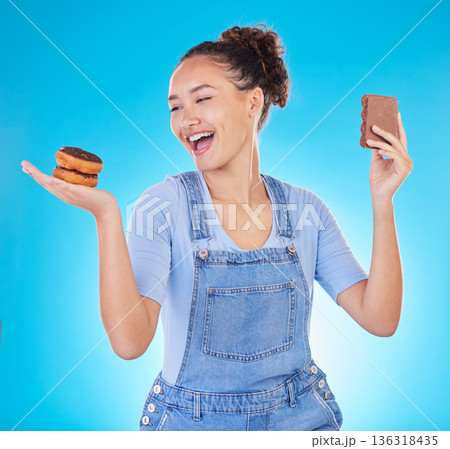 Food, diet and happy woman with sweets in studio, chocolate and donut for eating plan on blue background. Health, nutrition and choice to lose weight, girl with smile and freedom for sugar or dessert 136318435