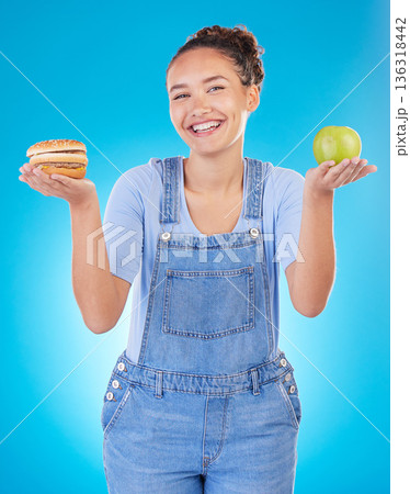 Happy, portrait and woman with burger, apple and choice in studio isolated on a blue background. Smile, fruit and person with sandwich, fast food or comparison for healthy diet, nutrition or wellness 136318442