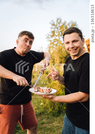 Happy friends serving grilled pork shashlik on a plate outdoors at sunset Happy friends serving grilled pork shashlik on a plate outdoors at sunset 136318613