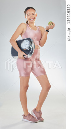 Portrait, scale and sports woman with apple on diet in studio for healthy results, weightloss goals and progress. Fitness, nutrition and happy female model lose weight with fruits on white background Portrait, scale and sports woman with apple on diet in studio for healthy results, weightloss goals and progress. Fitness, nutrition and happy female model lose weight with fruits on white background 136319520