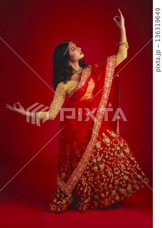 Fashion, dancer and Indian woman in a traditional dress, beauty and celebration on a red studio background. Female person, girl and model with a cultural outfit, dancing and confidence with religion Fashion, dancer and Indian woman in a traditional dress, beauty and celebration on a red studio background. Female person, girl and model with a cultural outfit, dancing and confidence with religion 136319649