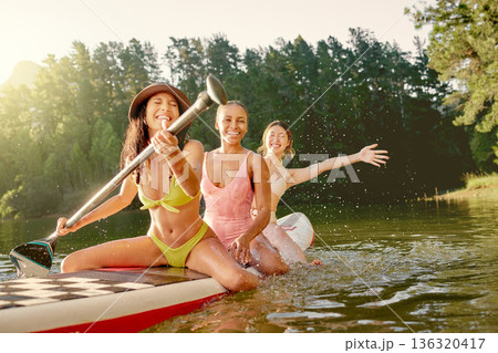 Girl friends, lake and paddle on board with smile, adventure and exercise in summer sunshine on holiday. Young women, water and together with rowing, excited and playing on vacation, river and forest 136320417