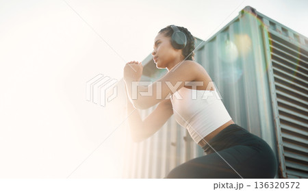 Fitness, woman with headphones and training with lens flare for motivation. Exercise or workout, health wellness or cardio and young female athlete squat in sportswear for healthy lifestyle 136320572