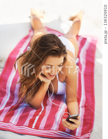 Happy woman, bikini and phone on beach for music in relax for audio listening or sound track outdoors. Female person lying on towel in swimwear enjoying listening to songs on mobile smartphone at sea Happy woman, bikini and phone on beach for music in relax for audio listening or sound track outdoors. Female person lying on towel in swimwear enjoying listening to songs on mobile smartphone at sea 136320910
