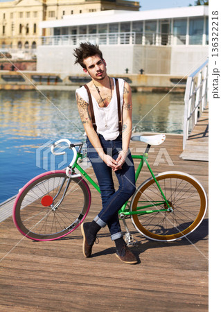 Bicycle, fashion and young man by the sea in a punk or rock outfit on an urban holiday. Tattoo, confidence and portrait of a serious grunge or edgy gen z male person with a bike by a lake boardwalk 136322218