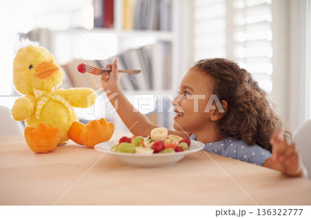 Child feeding, teddy bear and food at home with happiness and wellness meal with a smile. House, nutrition and young girl with healthy fruit salad and fruits of a hungry kid with toy duck at a table 136322777