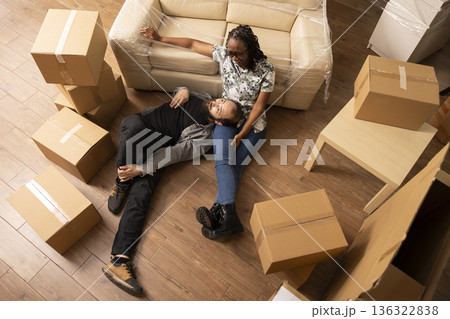 Caucasian boyfriend lying on lap of black girlfriend in living room filled with packed cardboard boxes. Diverse couple sits on floor, taking break during relocation day while waiting for moving truck. Caucasian boyfriend lying on lap of black girlfriend in living room filled with packed cardboard boxes. Diverse couple sits on floor, taking break during relocation day while waiting for moving truck. 136322838