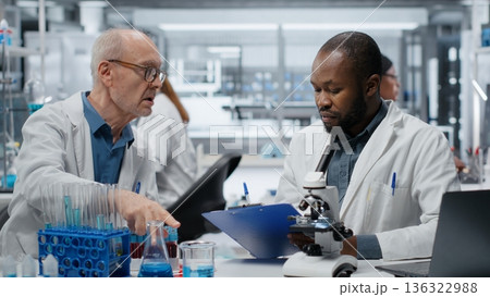 Medical team in lab conducting research on blood sample, taking notes. African american man and elderly colleague in laboratory analyzing sanguine fluid specimen, writing down findings, camera B 136322988
