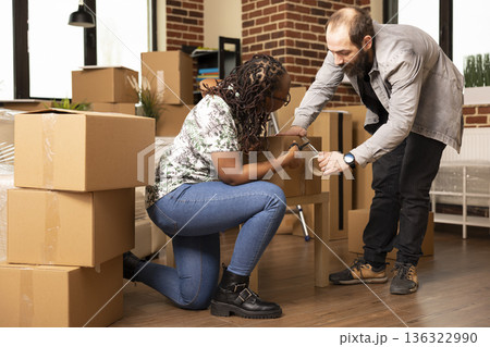 African american woman helping caucasian boyfriend by cutting adhesive tape while packing cardboard boxes. Young multiethnic couple carefully seals cartons, preparing to move out together. 136322990