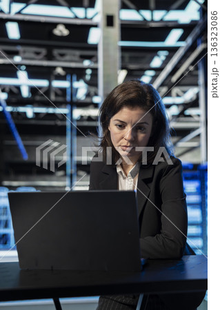 Data center female engineer using laptop to review equipment diagnostics. Worker at desk in server room using notebook to adjust configurations, applying firmware updates, cleaning error states Data center female engineer using laptop to review equipment diagnostics. Worker at desk in server room using notebook to adjust configurations, applying firmware updates, cleaning error states 136323086
