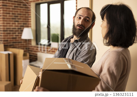 Caucasian husband and wife carrying belongings into brick wall apartment. White couple entering living room with moving boxes, chatting while relocating into newly purchased home. 136323111