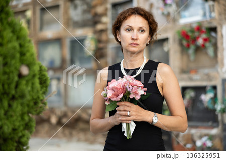 Sorrowful woman in black standing in cemetery with flowers 136325951