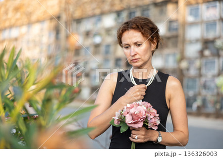 Sad woman in black dress with bouquet of flowers and handkerchief in hands, stands in cemetery 136326003