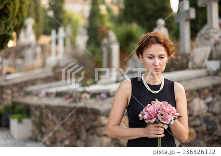 Sad woman in black dress with bouquet of flowers and handkerchief in hands, stands in cemetery 136326112