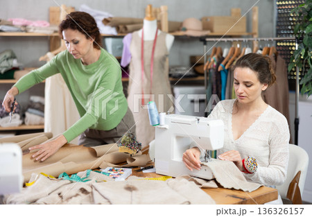 Female tailor sews on a sewing machine while another tailor cuts out patterns for dress with scissors 136326157