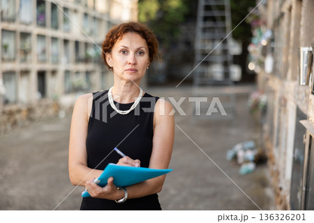 Female cemetery employee in black dress stands with report and writes down information Female cemetery employee in black dress stands with report and writes down information 136326201